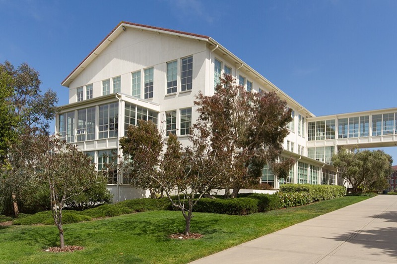 A white three-storey building with large windows and a footbridge, set in lawns and trees.